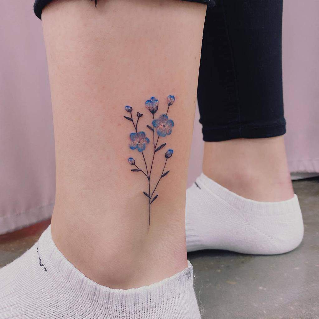 A delicate blue forget-me-not flower tattoo graces an ankle