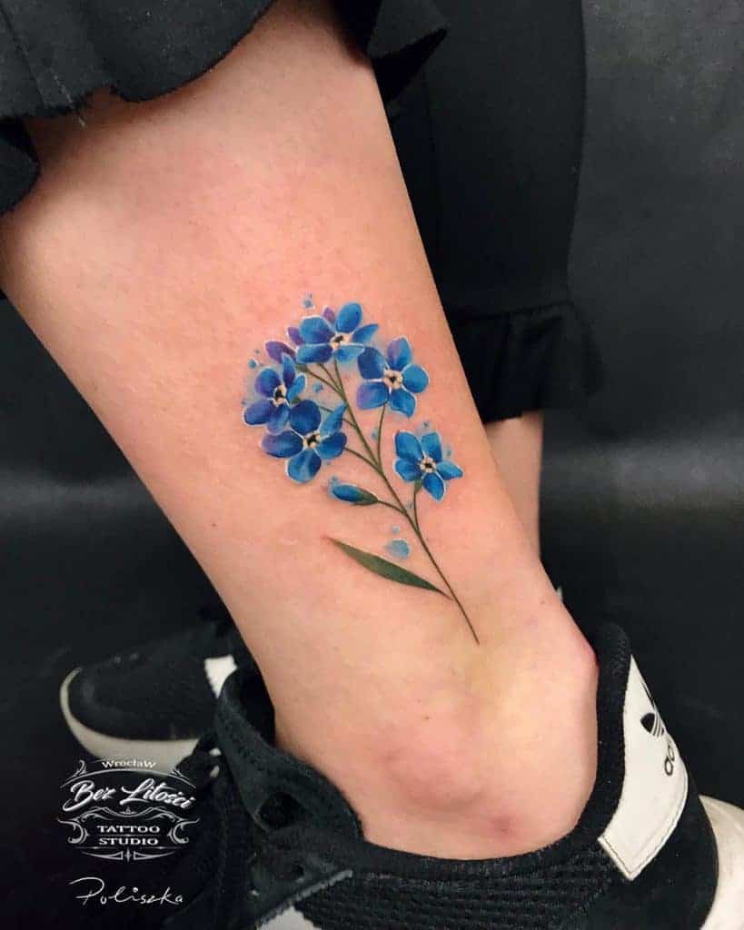 Delicate ankle tattoo features vibrant blue forget-me-not flowers, creating a stunningly detailed and elegant design