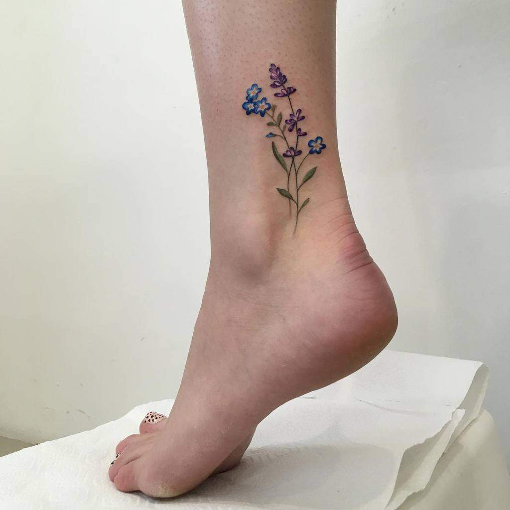 A delicate ankle tattoo featuring a cluster of forget-me-not flowers in shades of blue and purple, with soft green leaves, creating a gentle and feminine design
