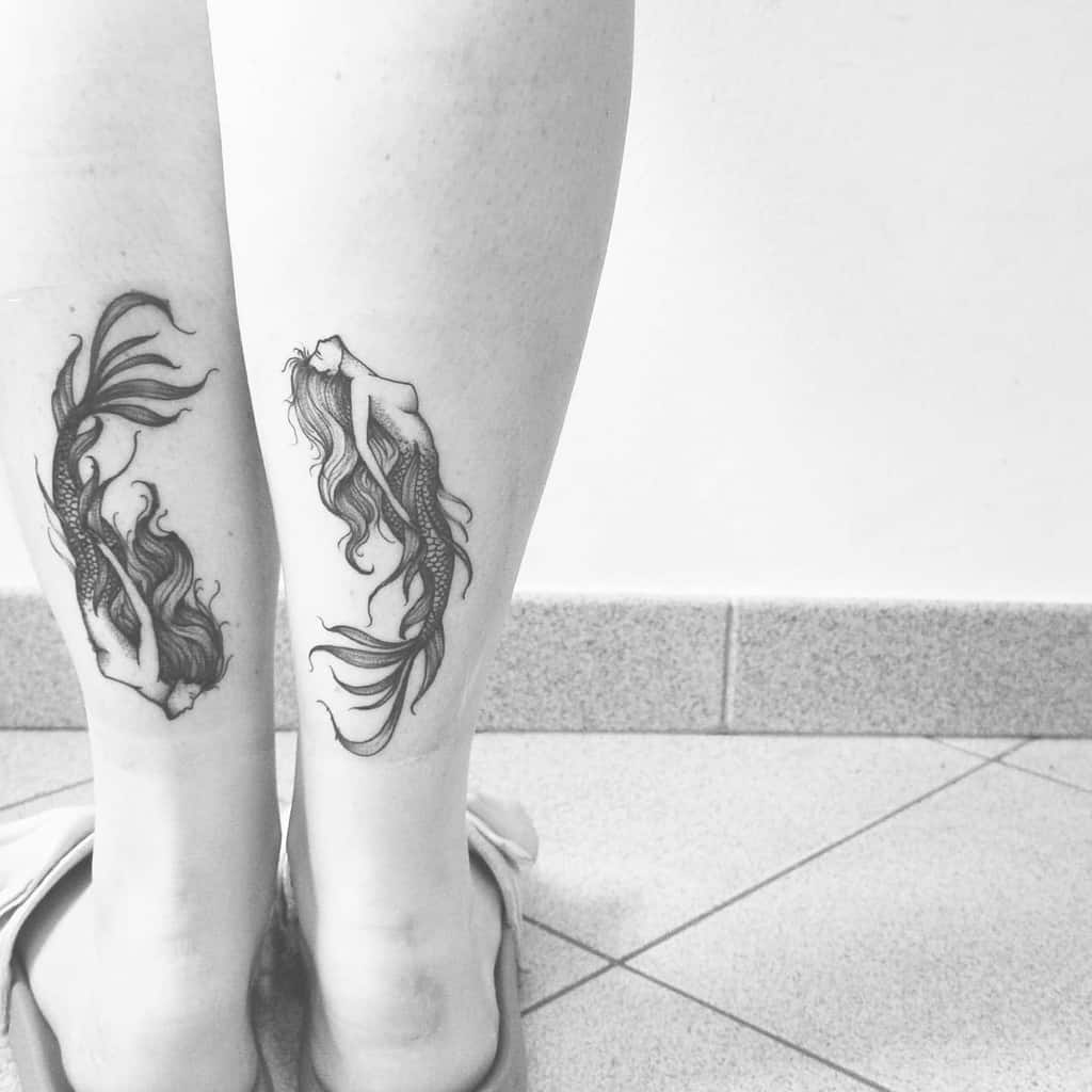 Matching black and grey mermaid tattoos on both legs, featuring flowing hair and delicate details in an elegant, minimalistic design.