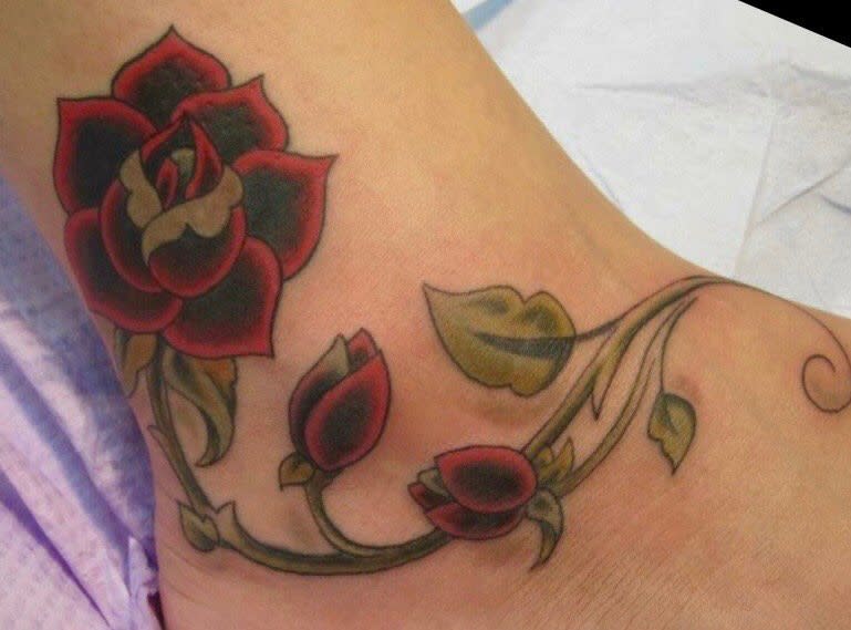 Elegant ankle tattoo featuring a bold red rose with budding flowers and flowing green vines, creating a graceful design