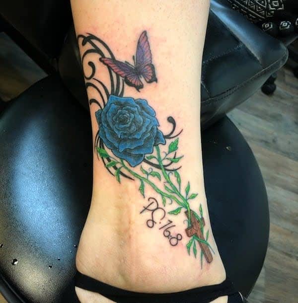 Vivid ankle tattoo featuring a striking blue rose intertwined with green vines, accompanied by a purple butterfly and a script text detail