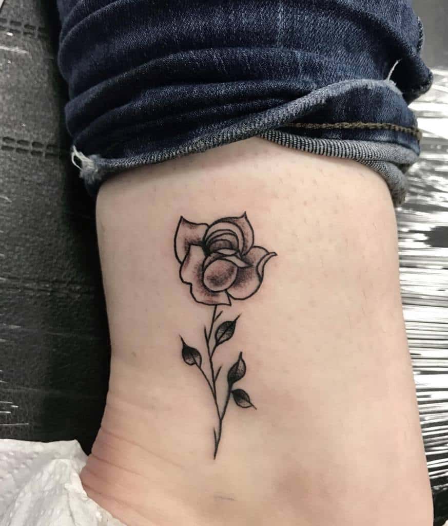 Black and grey rose tattoo on a person's ankle, with leaves and a stem stretching upward