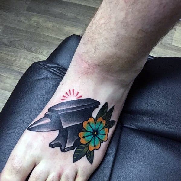 Anvil tattoo on foot featuring bold black ink, accented with a colorful orange and teal flower and green leaves.