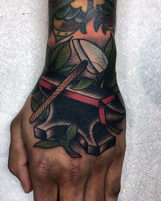 Anvil tattoo on the hand with a hammer resting on top, surrounded by green leaves and red ribbon in a bold traditional style.