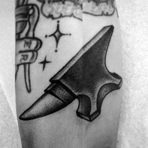 Black and grey anvil tattoo with bold shading, placed next to star symbols and part of a hand tattoo holding a tool.
