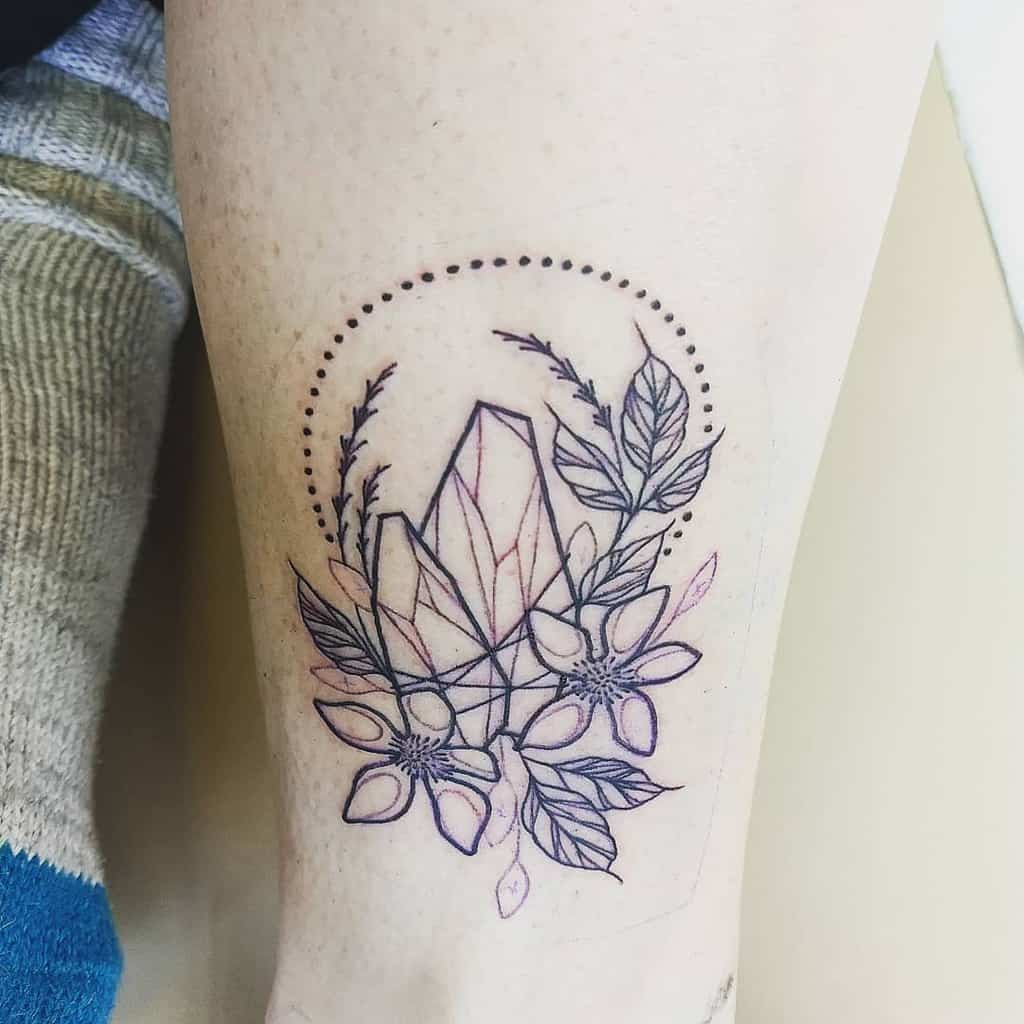 A geometric tattoo featuring a crystal surrounded by floral elements and leaves, with a dotted circle framing the design, placed on the arm.