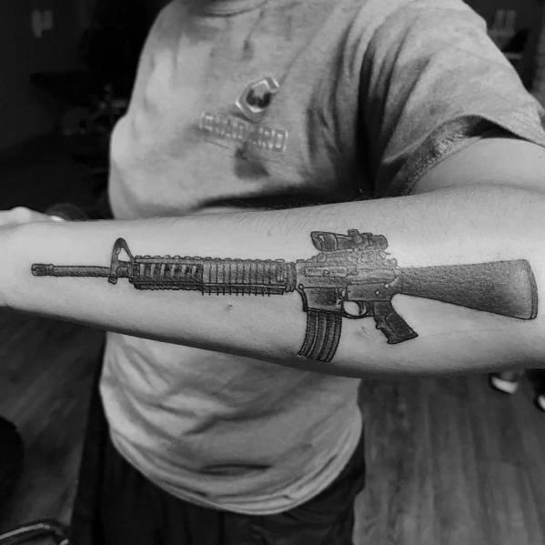 Detailed black and grey AR-15 tattoo on the forearm, featuring a scope and magazine, symbolizing tactical precision and firearm appreciation.