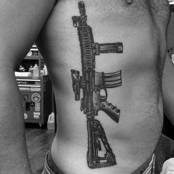 Large black and grey AR-15 tattoo on the ribcage, featuring intricate details, a tactical grip, and a suppressor, symbolizing strength and precision.