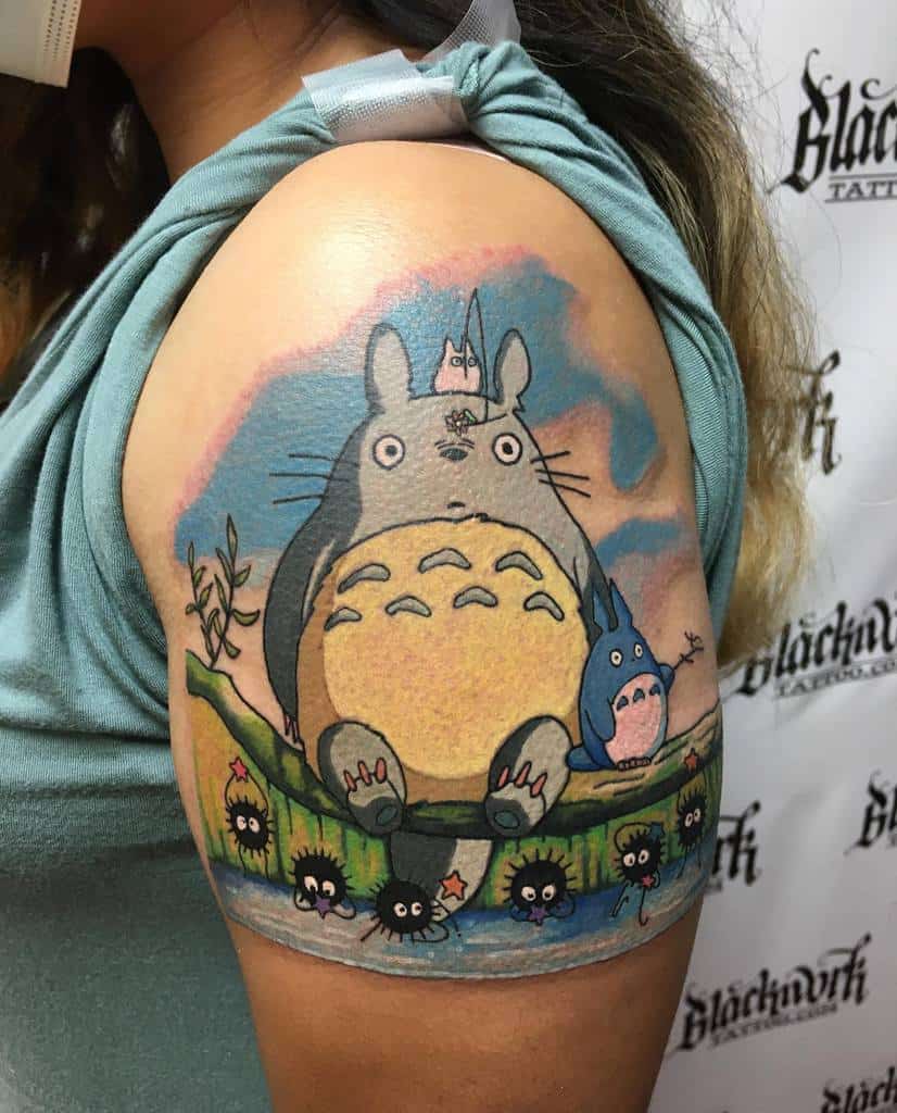 Vibrant upper arm tattoo of Totoro sitting by a pond with soot sprites, featuring detailed colors and a scenic background