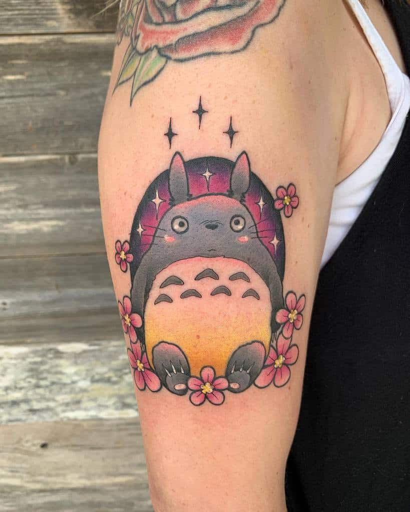 Colorful Totoro tattoo on the upper arm, surrounded by pink flowers and sparkles, with a sunset gradient background