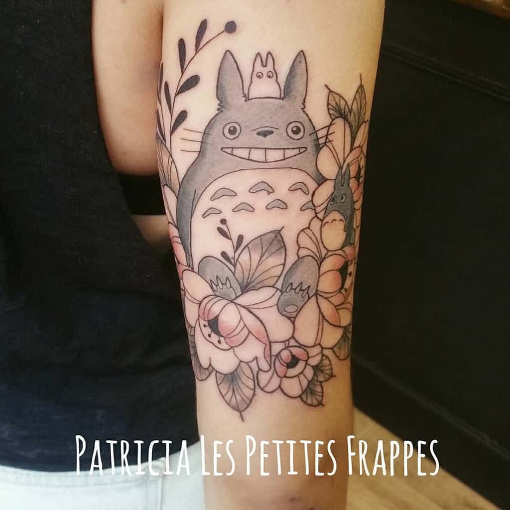 Black and grey Totoro tattoo on the arm with floral details and leaves, featuring Totoro and smaller companions