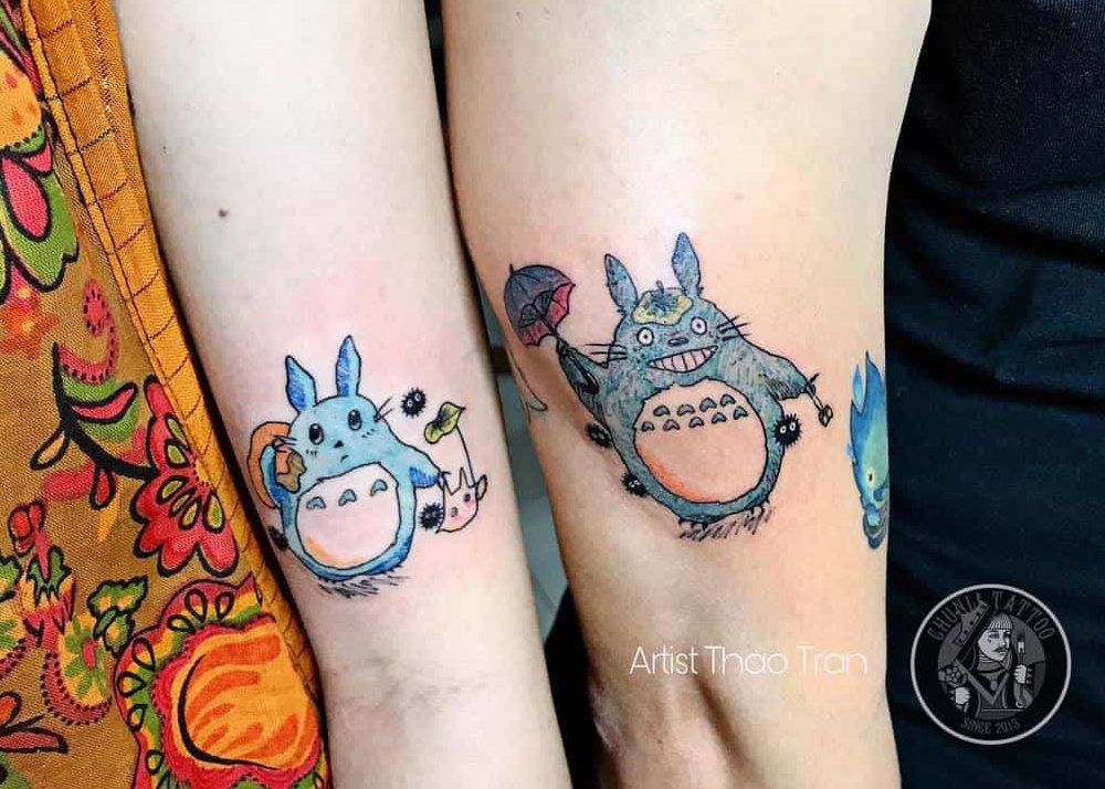 Matching Totoro tattoos on two arms, featuring colorful Totoros with soot sprites, leaves, and umbrellas