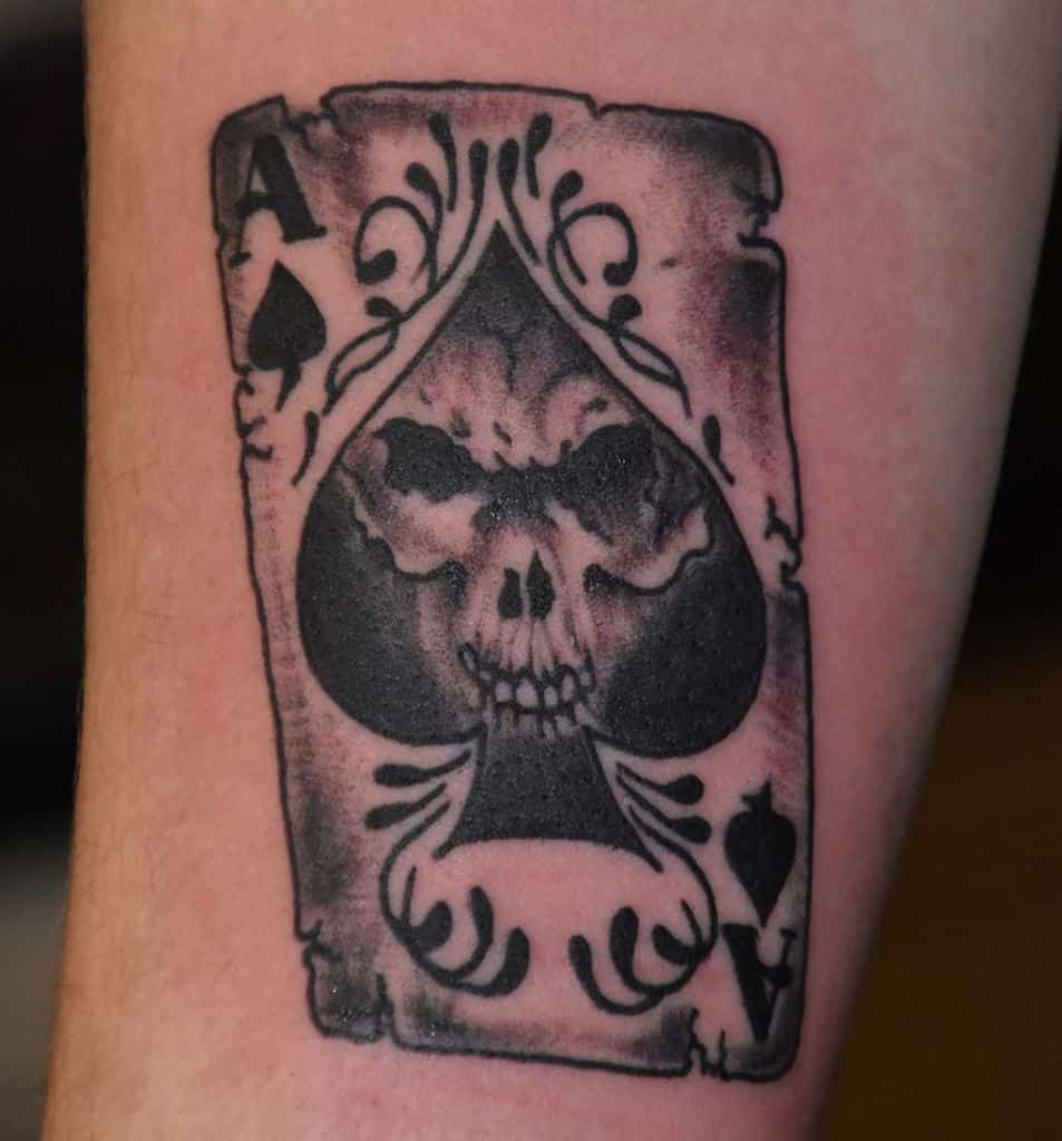 A black-and-white tattoo of the Ace of Spades card with a skull in the center, framed by ornate detailing and shading