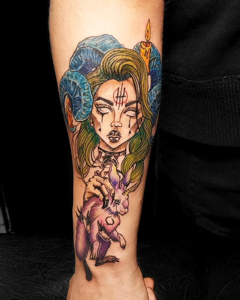A tattoo inspired by Alice in Wonderland, featuring a woman with ram horns holding a purple rabbit, candles, and a tear-streaked face