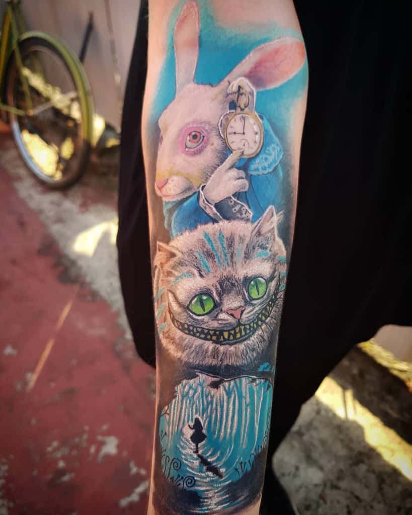 A colorful tattoo featuring the White Rabbit holding a pocket watch and the Cheshire Cat from Alice in Wonderland and Alice exploring the forest below