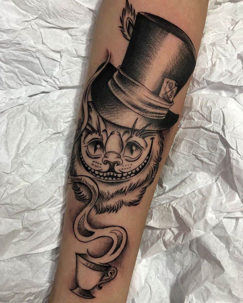 A black and grey tattoo of the Cheshire Cat from Alice in Wonderland, featuring a teacup, steam, and the Mad Hatter's hat