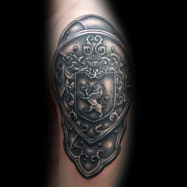 Intricate upper arm tattoo of an ornamental shield with a lion crest in the center, detailed with scrollwork and metallic shading.