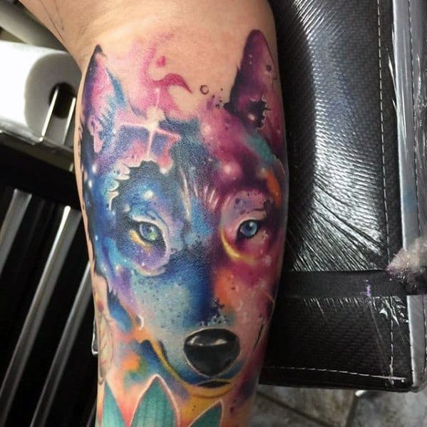Wolf tattoo in vibrant watercolor style with cosmic and abstract elements blending purples, blues, and pinks on forearm.