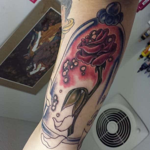 A vibrant Beauty and the Beast rose tattoo inside a glass dome with deep red tones, soft shading, and scattered petals