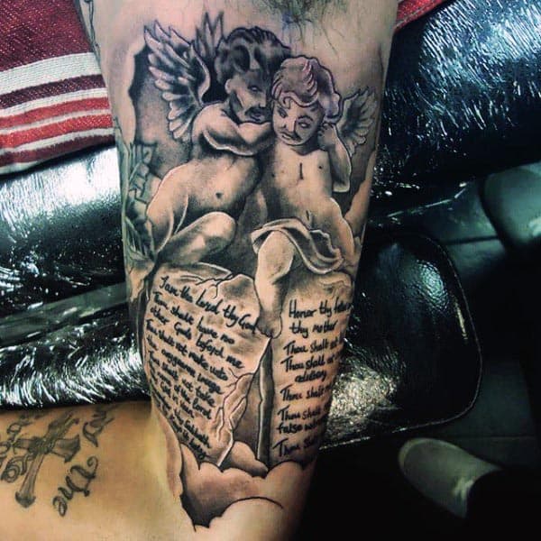Black and grey tattoo of two cherubs holding stone tablets with engraved biblical text, featuring detailed shading and realism