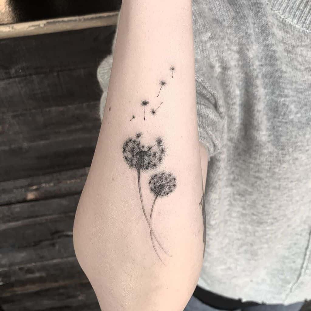 Fine-line dandelion tattoo on the forearm with two flowers and seeds blowing away, symbolizing growth, freedom, and wishes.