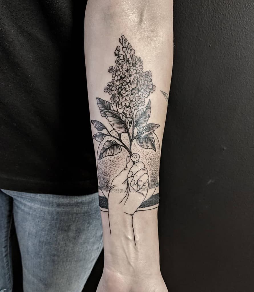 Black and grey tattoo of a hand holding a lilac branch, featuring fine-line details and dot shading on the forearm.