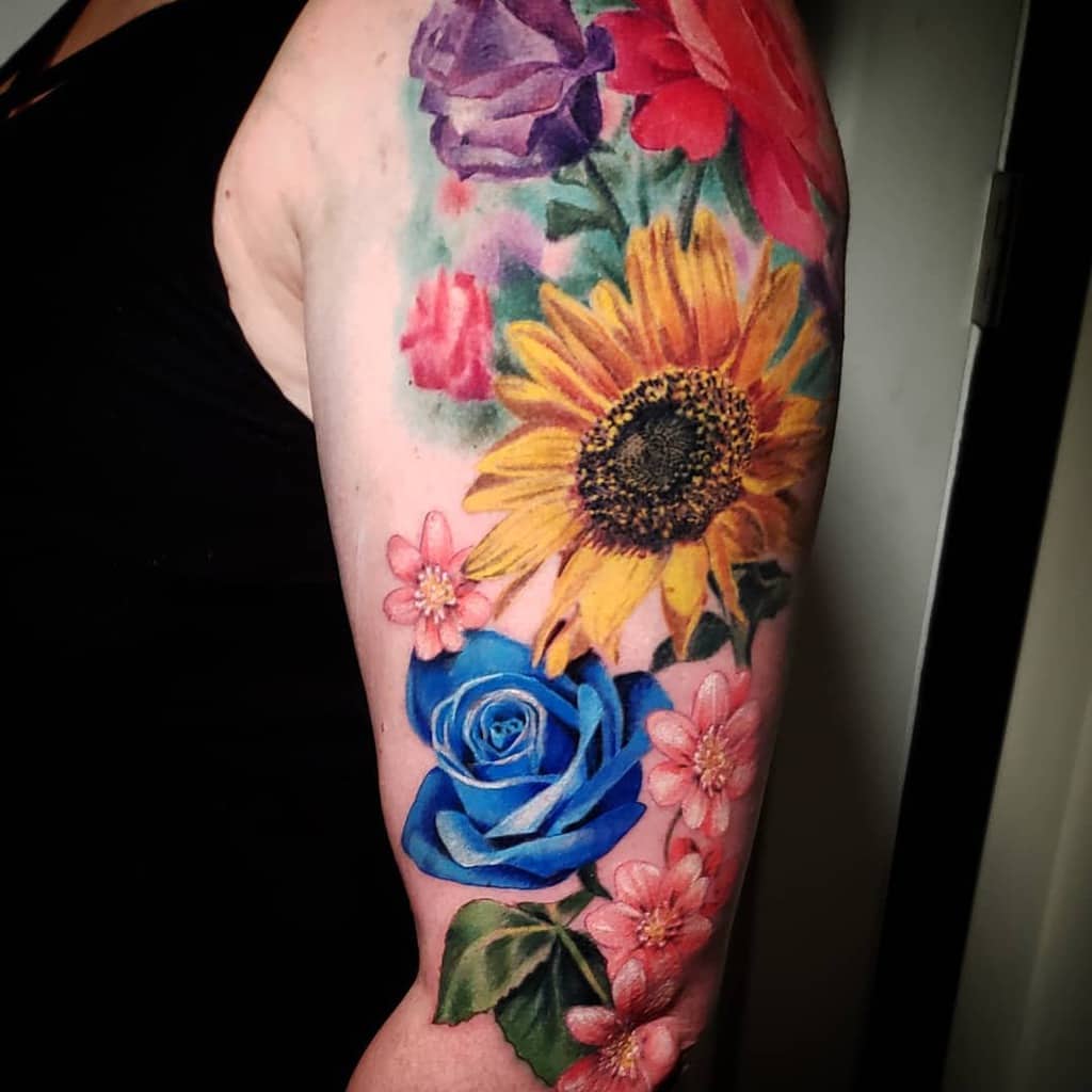 An arm tattoo showcasing vibrant flowers, with a striking blue rose set amidst pink daisies and a radiant sunflower