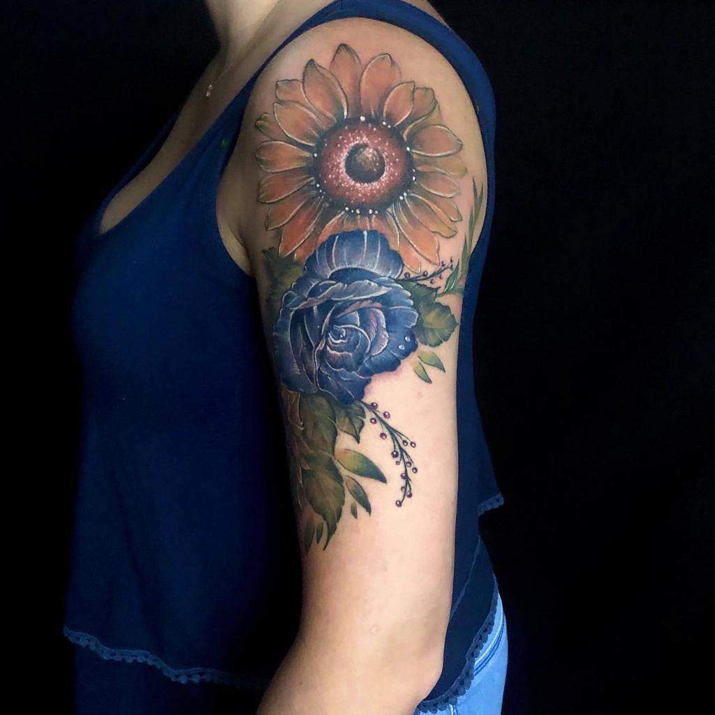 A vibrant arm tattoo featuring a sunflower, intricate blue rose, and lush leaves