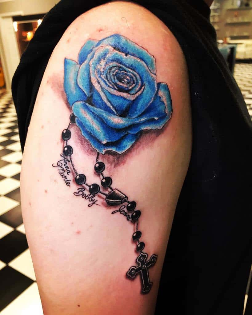 A captivating tattoo of a blue rose entwined with a rosary adorns the person's upper arm, names gracefully inscribed within its design