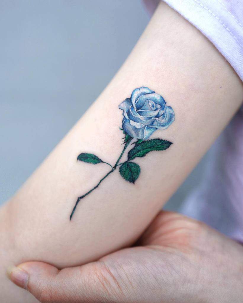 Stunning blue rose tattoo elegantly etched on an arm, complete with its delicate stem and vibrant green leaves