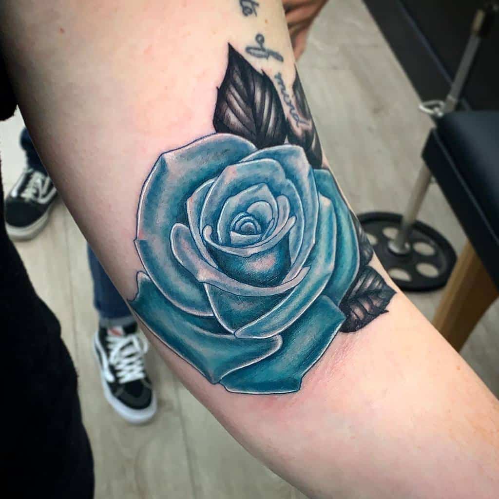 A realistic blue rose tattoo on the forearm, showcasing intricate shading and highlights to create depth, with dark leaves in the background