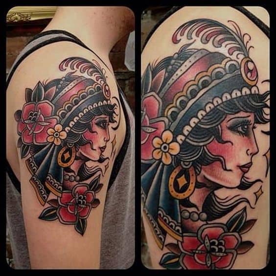 Bold traditional gypsy woman tattoo on upper arm, featuring vibrant red flowers, gold horseshoe earring, and detailed shading.