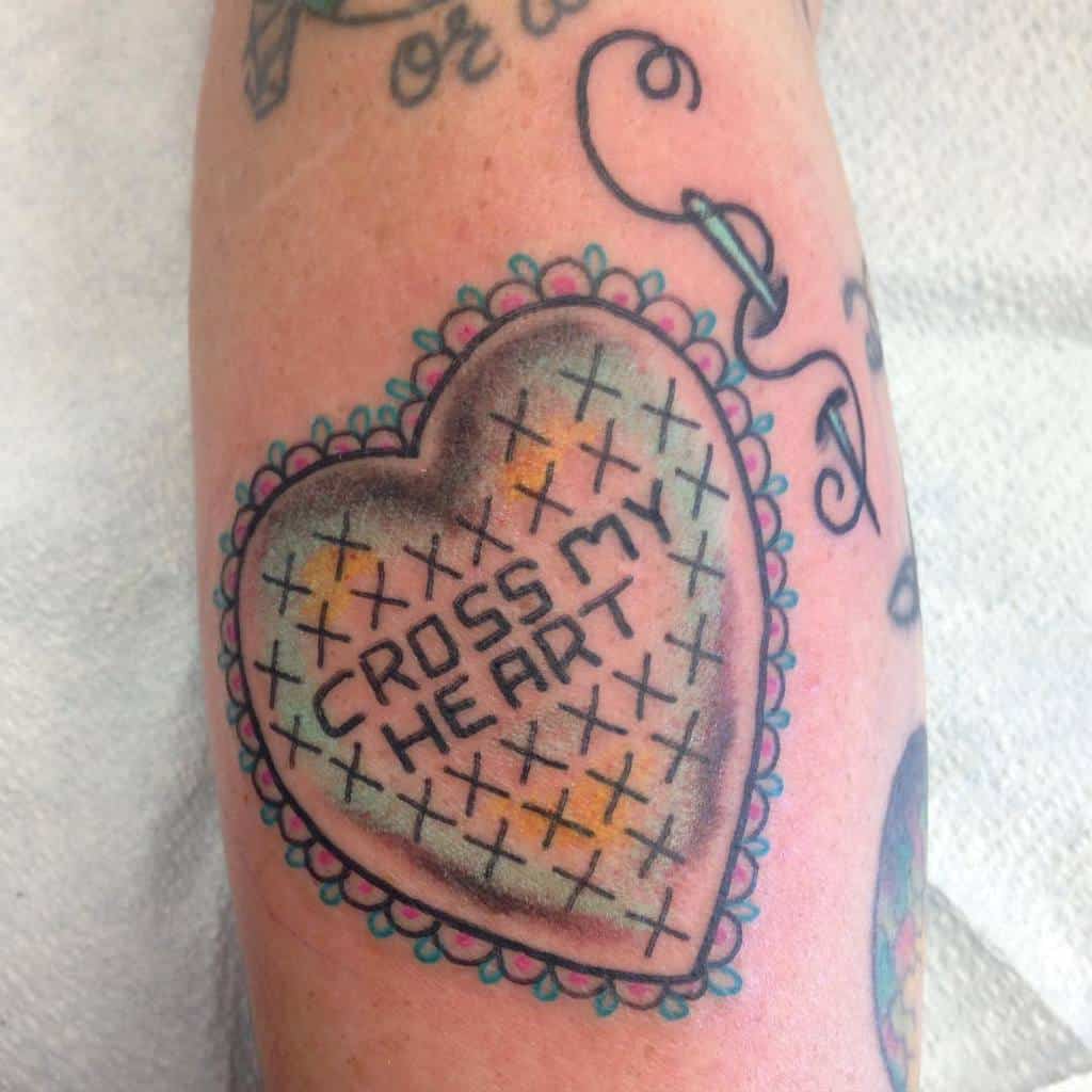 Cross-stitch tattoo of a heart-shaped patch with 'CROSS MY HEART' text, bordered with decorative stitching and a threaded needle design