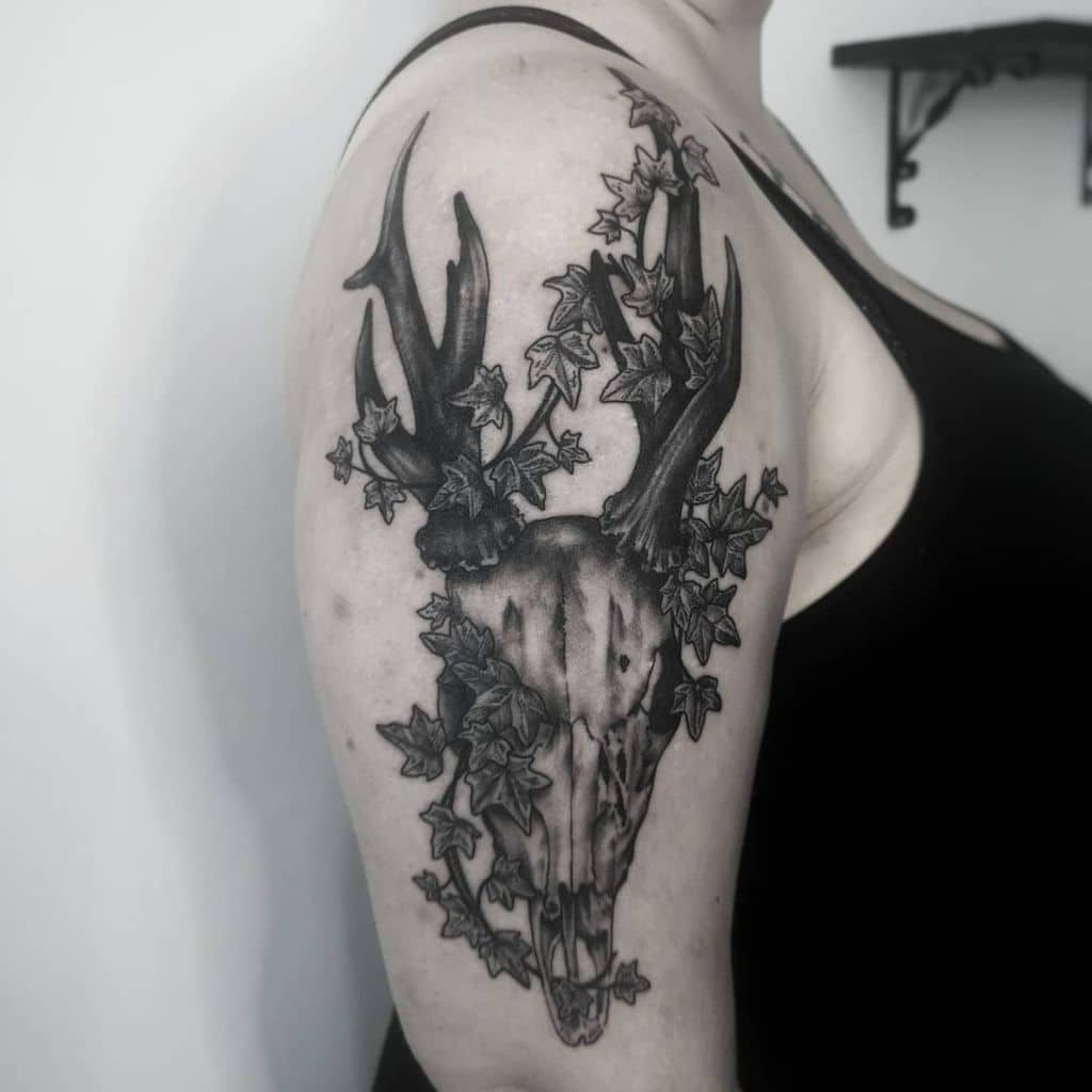 Black and grey tattoo of a deer skull entwined with ivy leaves on a person's upper arm