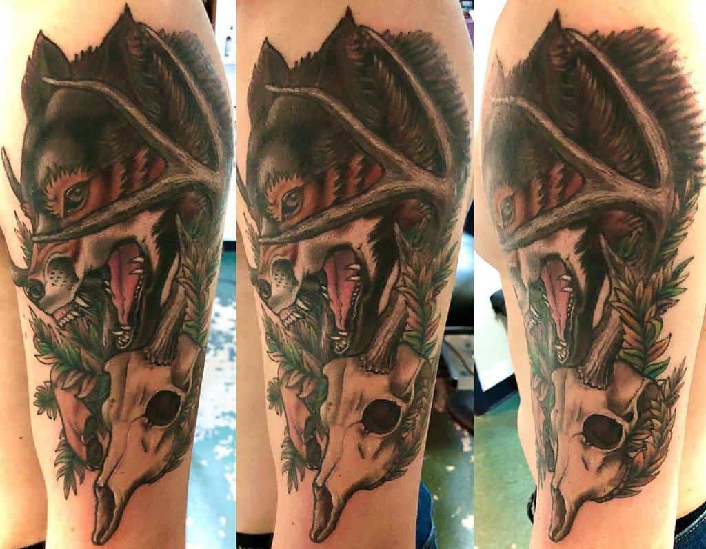 Tattoo of a snarling wolf with antlers and a deer skull, surrounded by green foliage