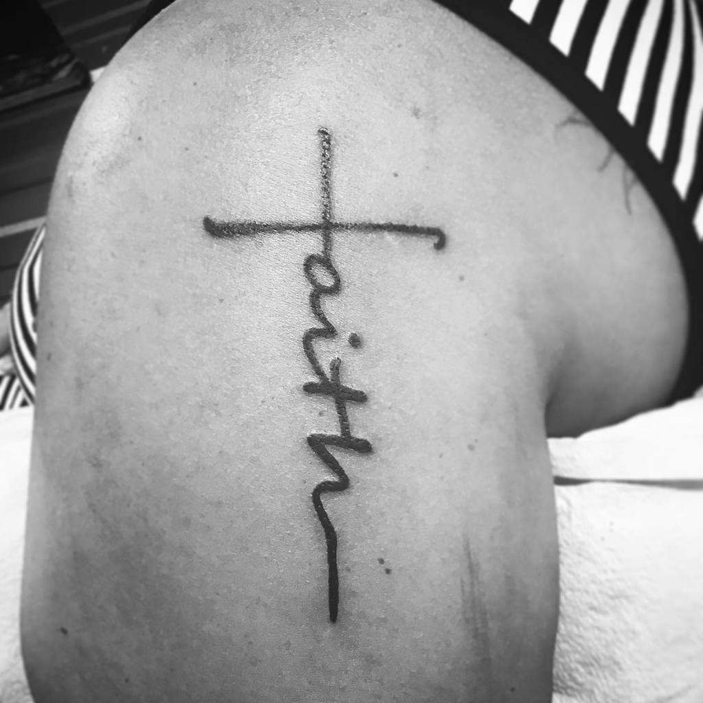 A tattoo elegantly weaves the word "faith" into a cross shape on an arm, symbolizing unwavering belief and strength
