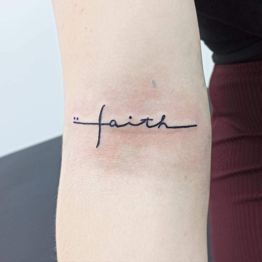 A minimalist cross and 'faith' tattoo in black ink on the inner arm