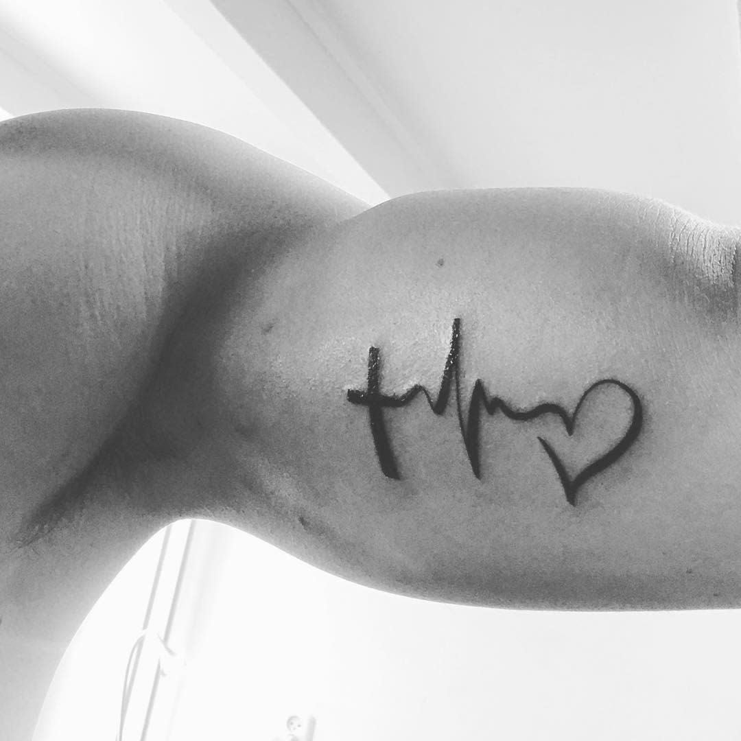 Black and white photo of a bicep tattoo elegantly intertwining a cross, heartbeat line, and heart, symbolizing faith, hope and love