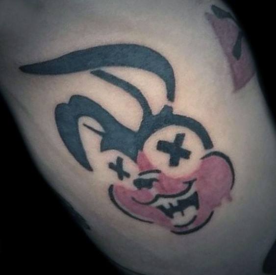 Tattoo of a twisted cartoon rabbit with black X eyes, red cheeks, and a mischievous grin in a bold, punk style.
