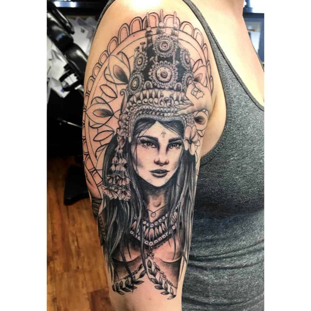 A detailed black and gray tattoo of a woman with a headdress, feathers, and intricate designs on the upper arm