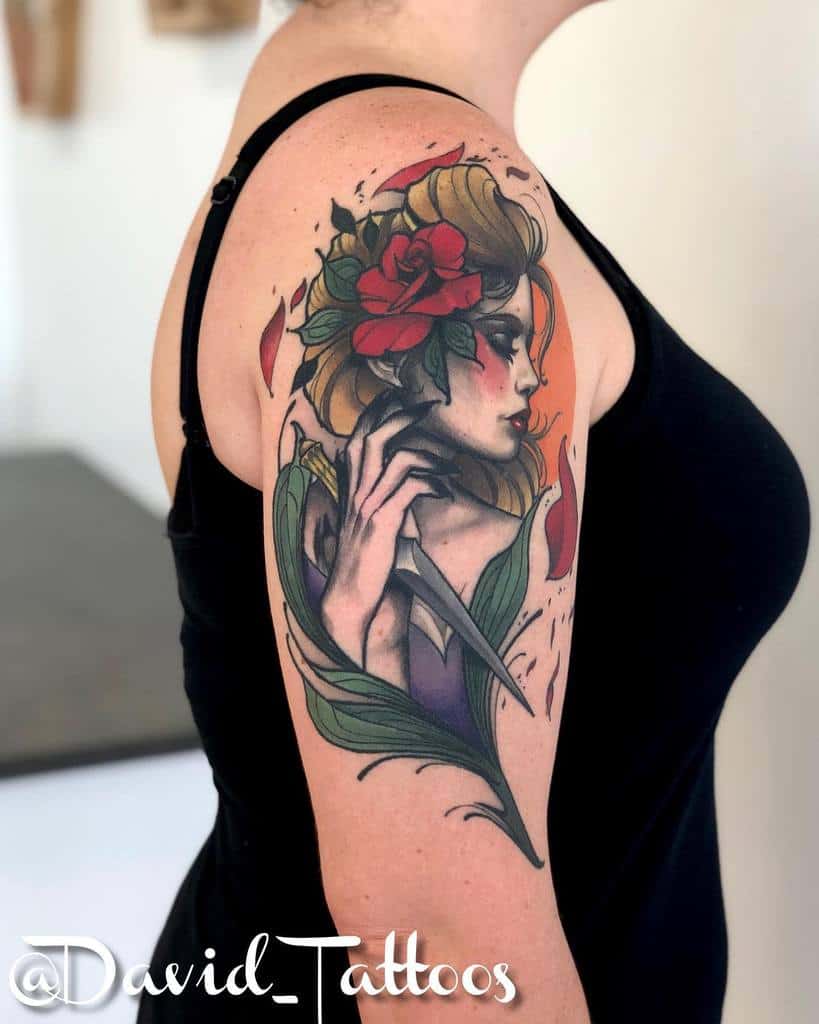 A vibrant tattoo of a gypsy woman with a rose in her hair, holding a knife, surrounded by flowing leaves, on the upper arm