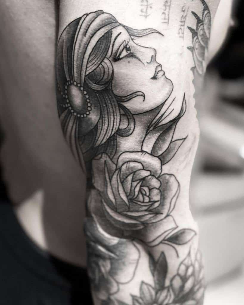 Gypsy-inspired tattoo of a woman's face intertwined with roses on an arm, crafted in black and gray ink