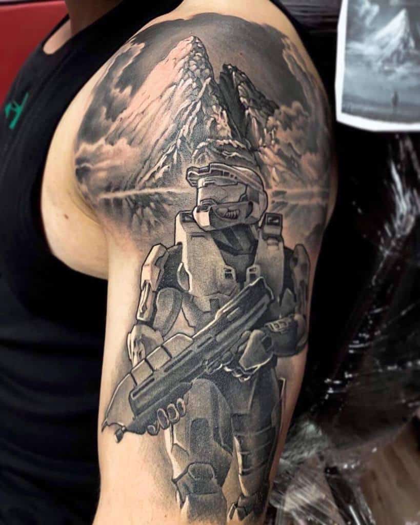 Epic black and gray Halo tattoo of Master Chief standing with a shotgun, set against a detailed mountain landscape, inked as a shoulder piece.