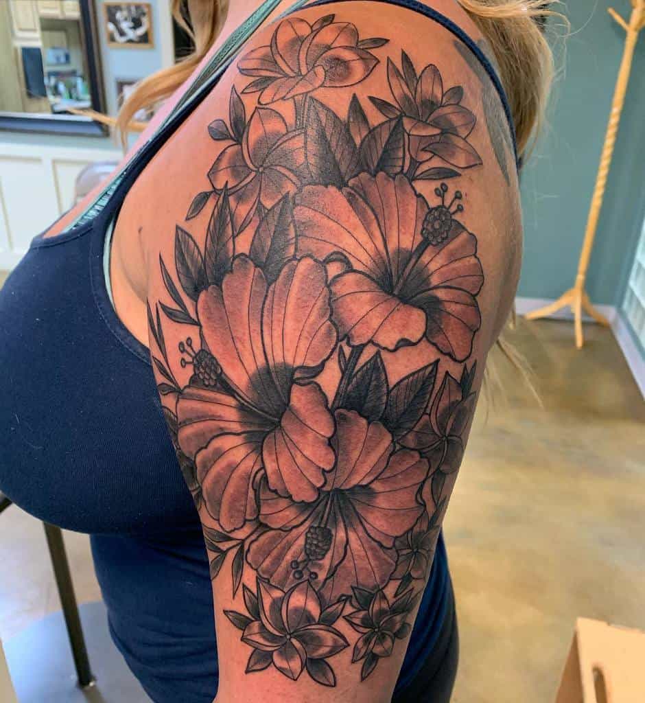 Upper arm tattoo featuring detailed black and grey hibiscus and plumeria flowers with intricate shading and leafy accents