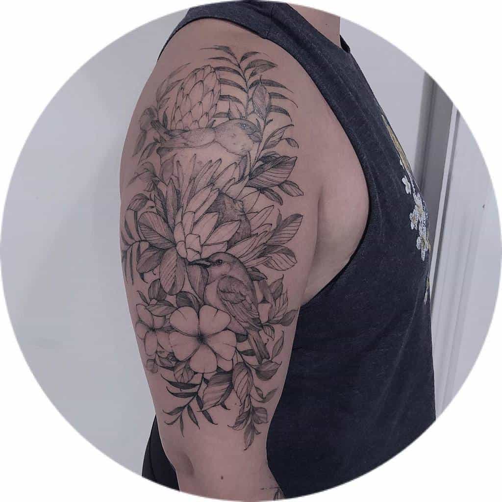 Upper arm tattoo featuring detailed black and grey plumeria flowers, lush leaves, and two birds, creating a natural, tropical design