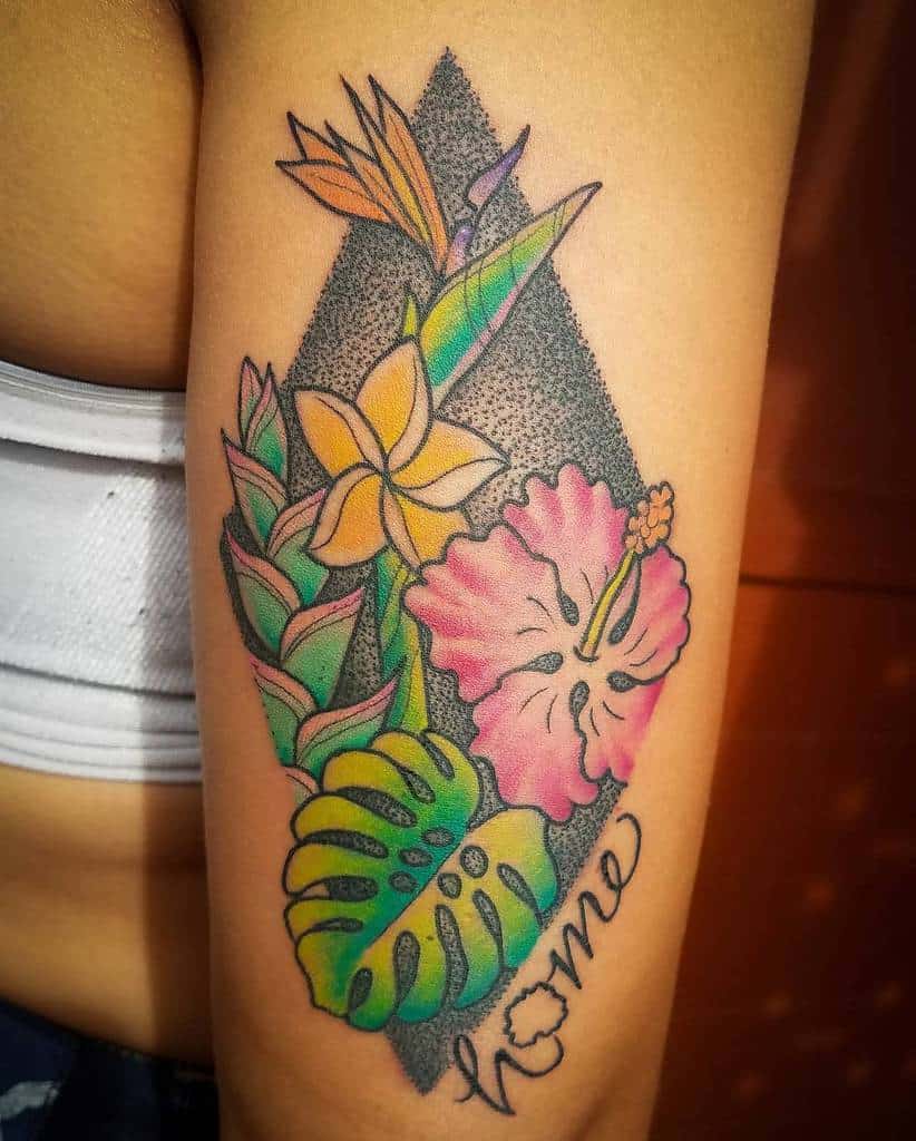Upper arm tattoo featuring a yellow plumeria, pink hibiscus, bird of paradise flower, and tropical leaves with the word 'home' scripted