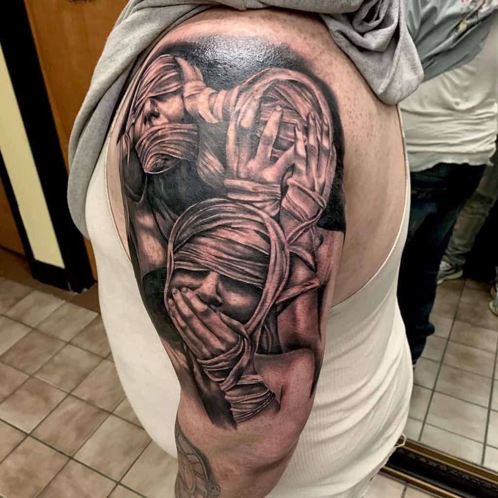 Black and gray tattoo of figures with bandaged eyes and mouths, covering ears, on an upper arm