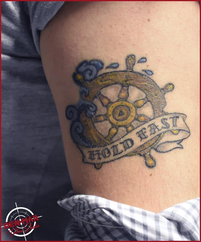 Tattoo of a ship's wheel with "Hold Fast" banner on an arm