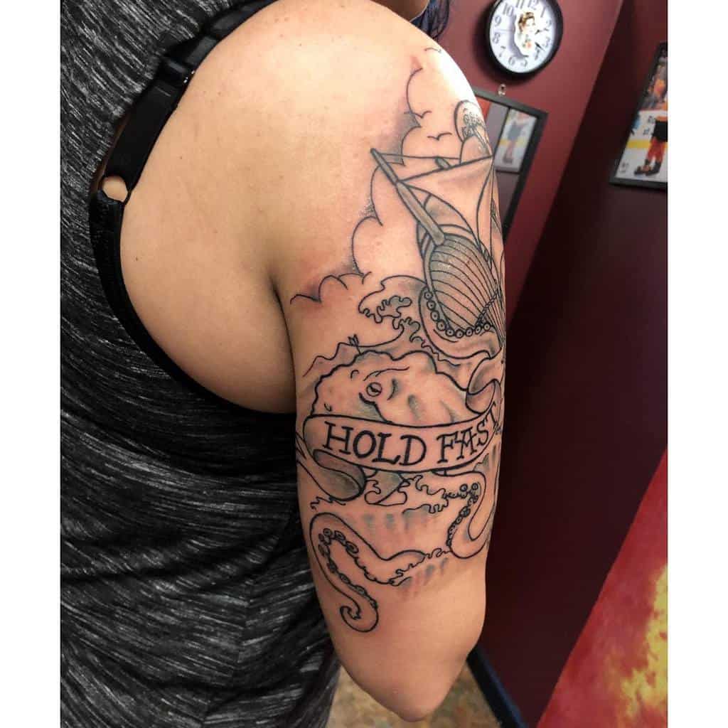 Tattoo of a kraken holding a ship with "Hold Fast" banner on upper arm